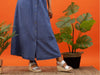 Lunar Rochester JLH507-Grey metallic wedge sandals with double straps and buckles, resting on a woven platform, worn by a lady wearing a maxi denim button down dress. Two large green plants on either side of her against an orange background and on a woven mat. 