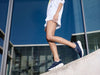 Stylish white Tenhaag 0001-057 Navy sports shoes worn by a person standing on a slanting ledge with glass building in the background. The sneakers feature a modern design with breathable mesh panels, elasticated laces, and a cushioned sole with cut out details. The gold branding and sleek finish add a touch of elegance, making them perfect for casual and athleisure wear.